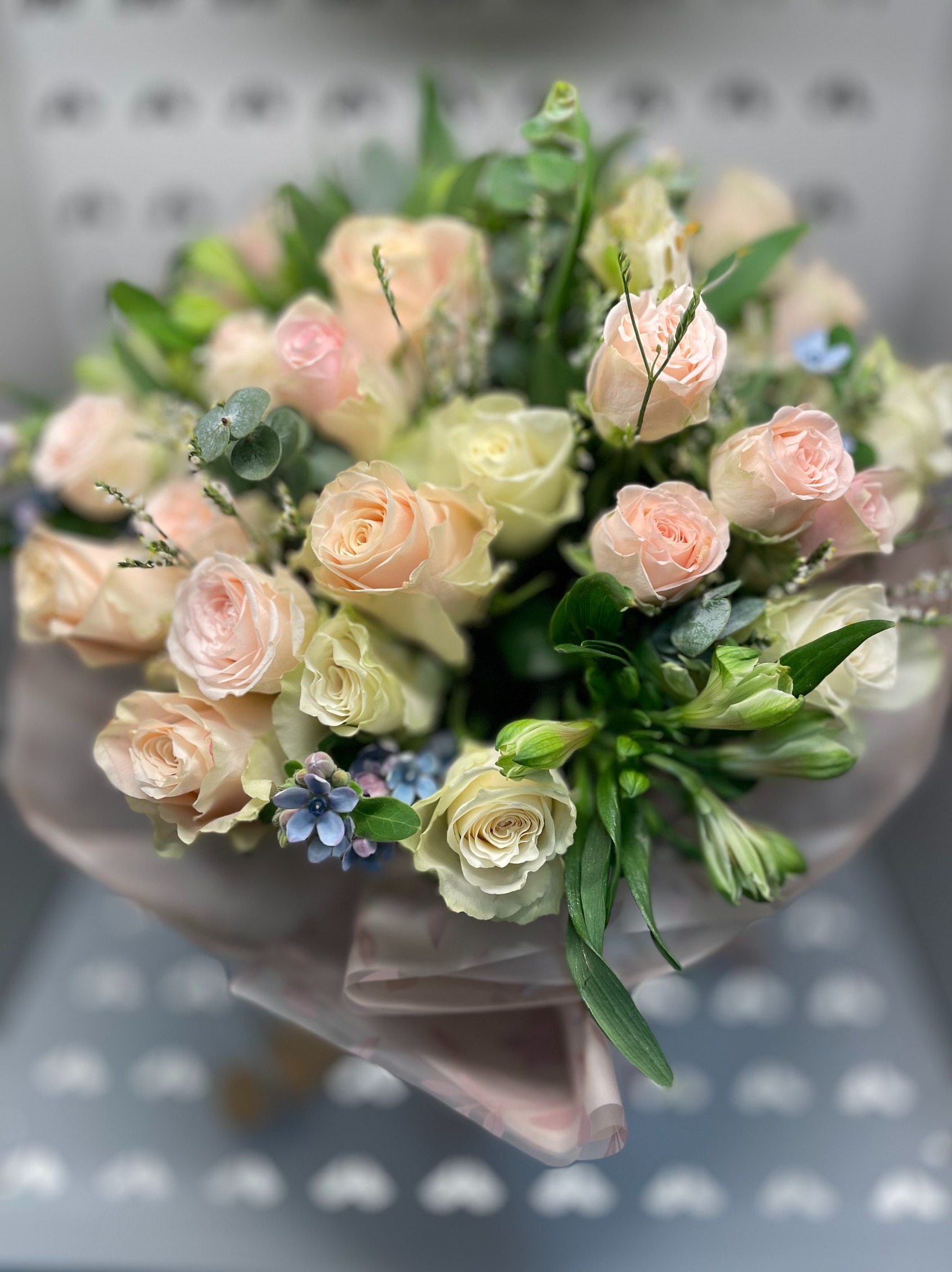 Delikatny bukiet z róż w odcieniach kremu i różu, uzupełniony zielenią i drobnymi niebieskimi kwiatami, zapakowany w ozdobny papier. Romantyczna kompozycja kwiatowa.