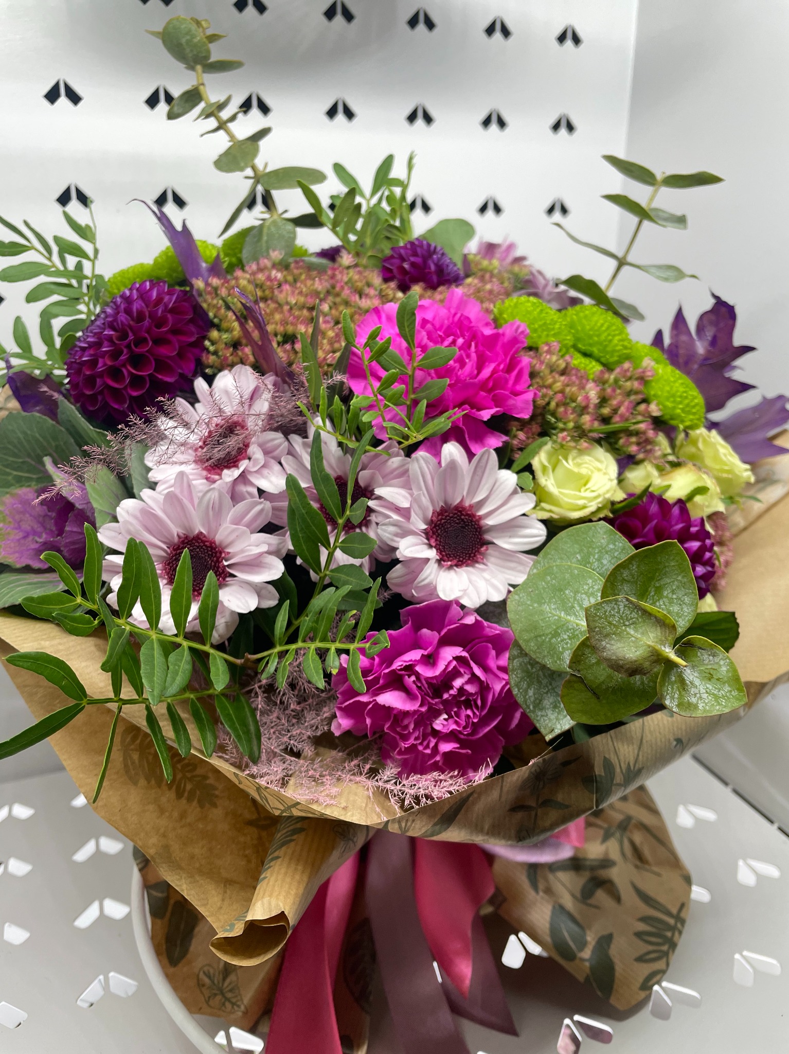Bukiet mieszany z różowych goździków, białych gerber i fioletowych dalii, owinięty w papier z motywem liści, przewiązany różową wstążką.