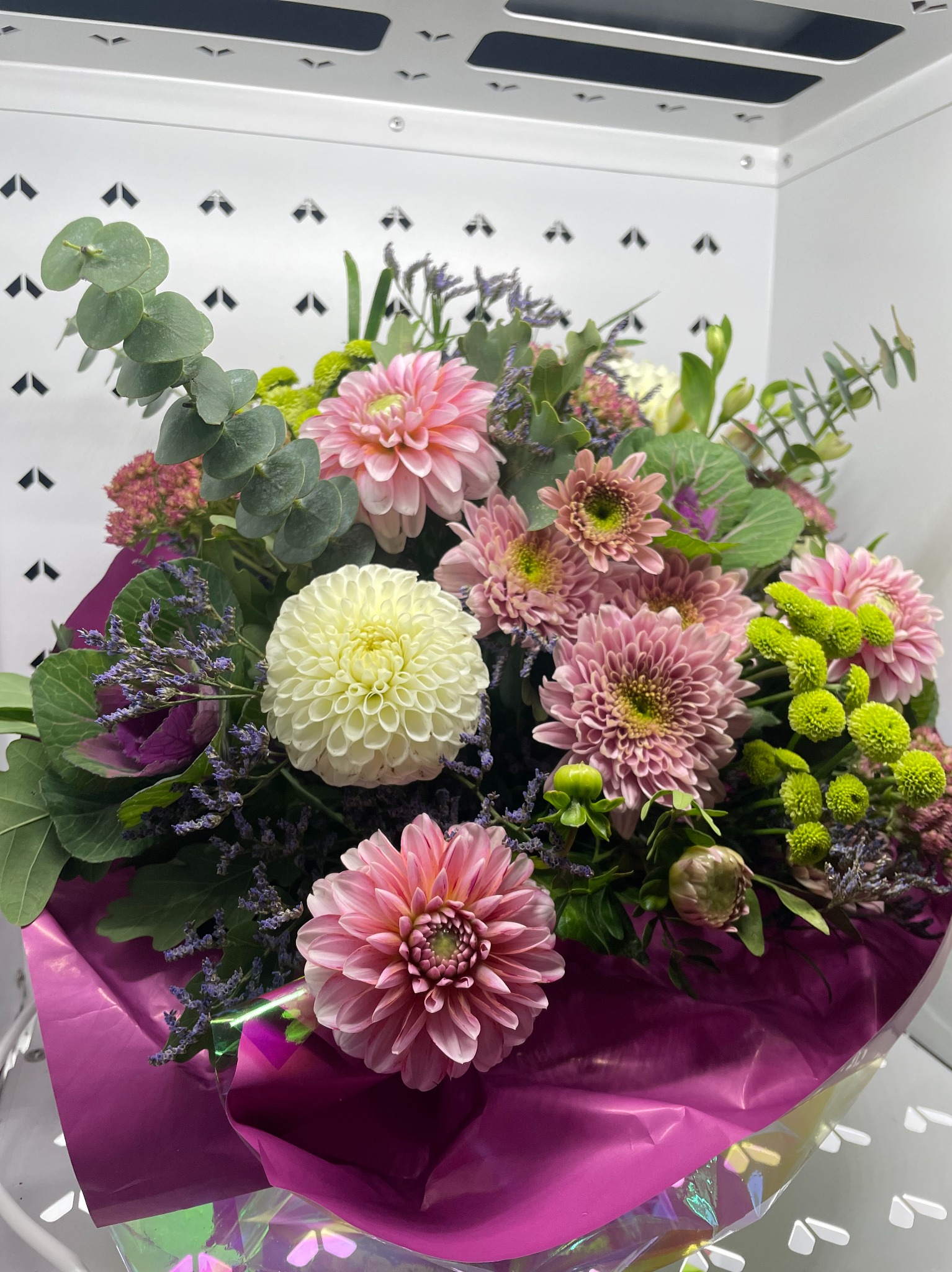 Bukiet mieszanych kwiatów w odcieniach różu, bieli i zieleni, owinięty w liliowy papier. Widoczny eukaliptus i drobne fioletowe kwiaty. Tło to biała ściana.