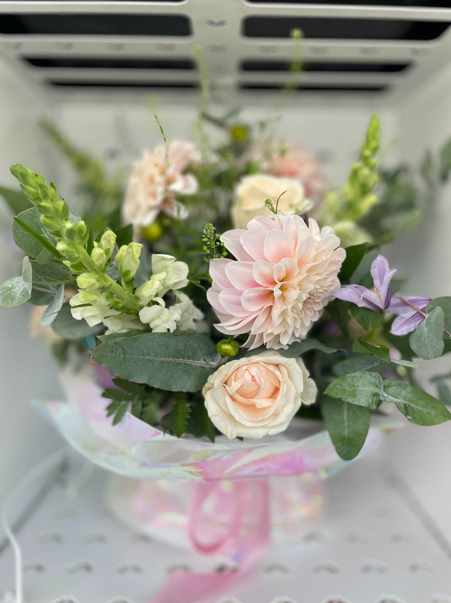 Delikatny bukiet z różowych dalii, kremowych róż i zielonych dodatków, zapakowany w ozdobne, opalizujące pudełko z różową wstążką. Romantyczna kompozycja kwiatowa.