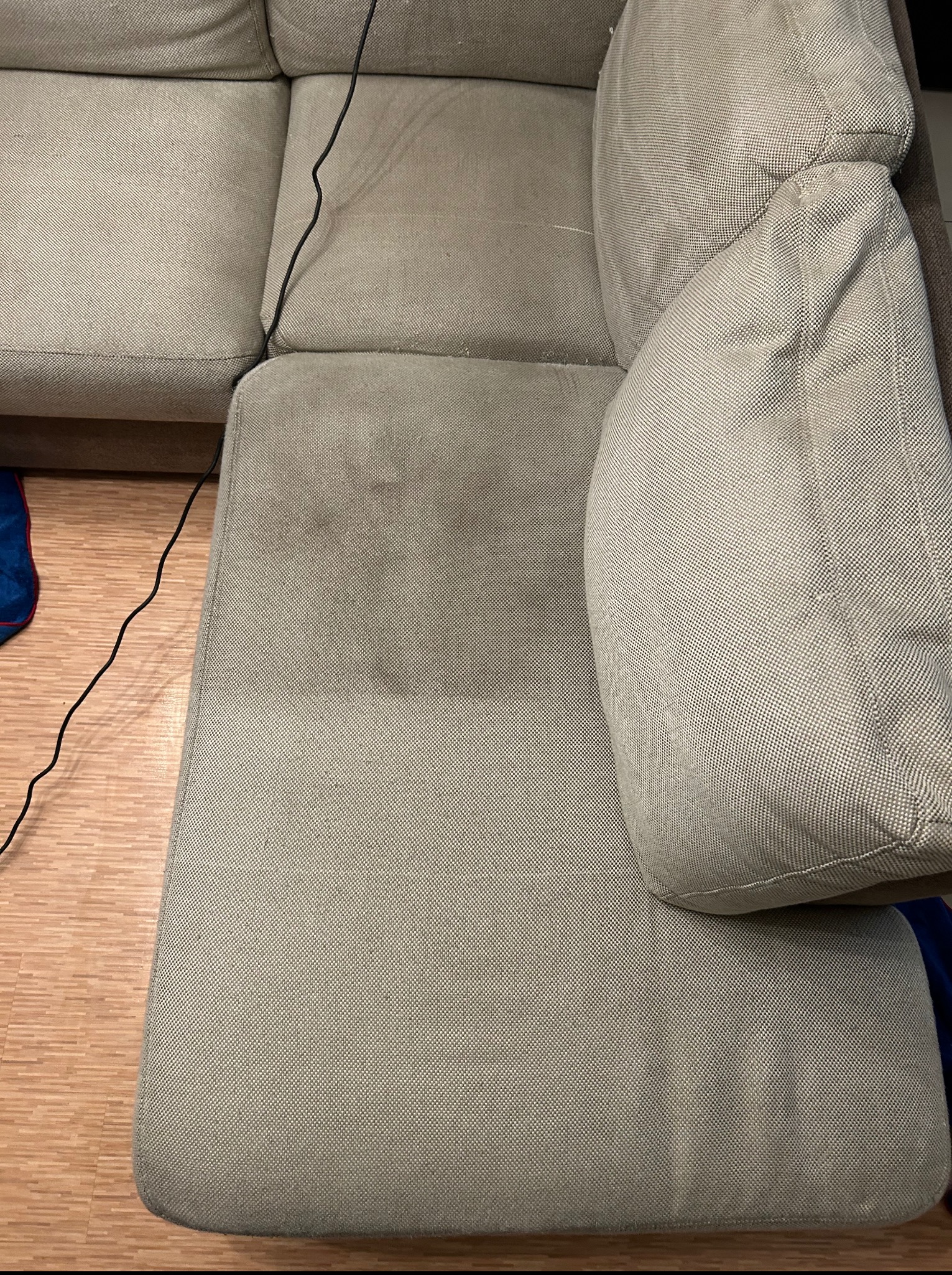 Częściowo wyczyszczona, beżowa kanapa z widocznym efektem prania tapicerki na siedzisku. Na podłodze widoczny fragment kabla i niebieskiej tkaniny.
