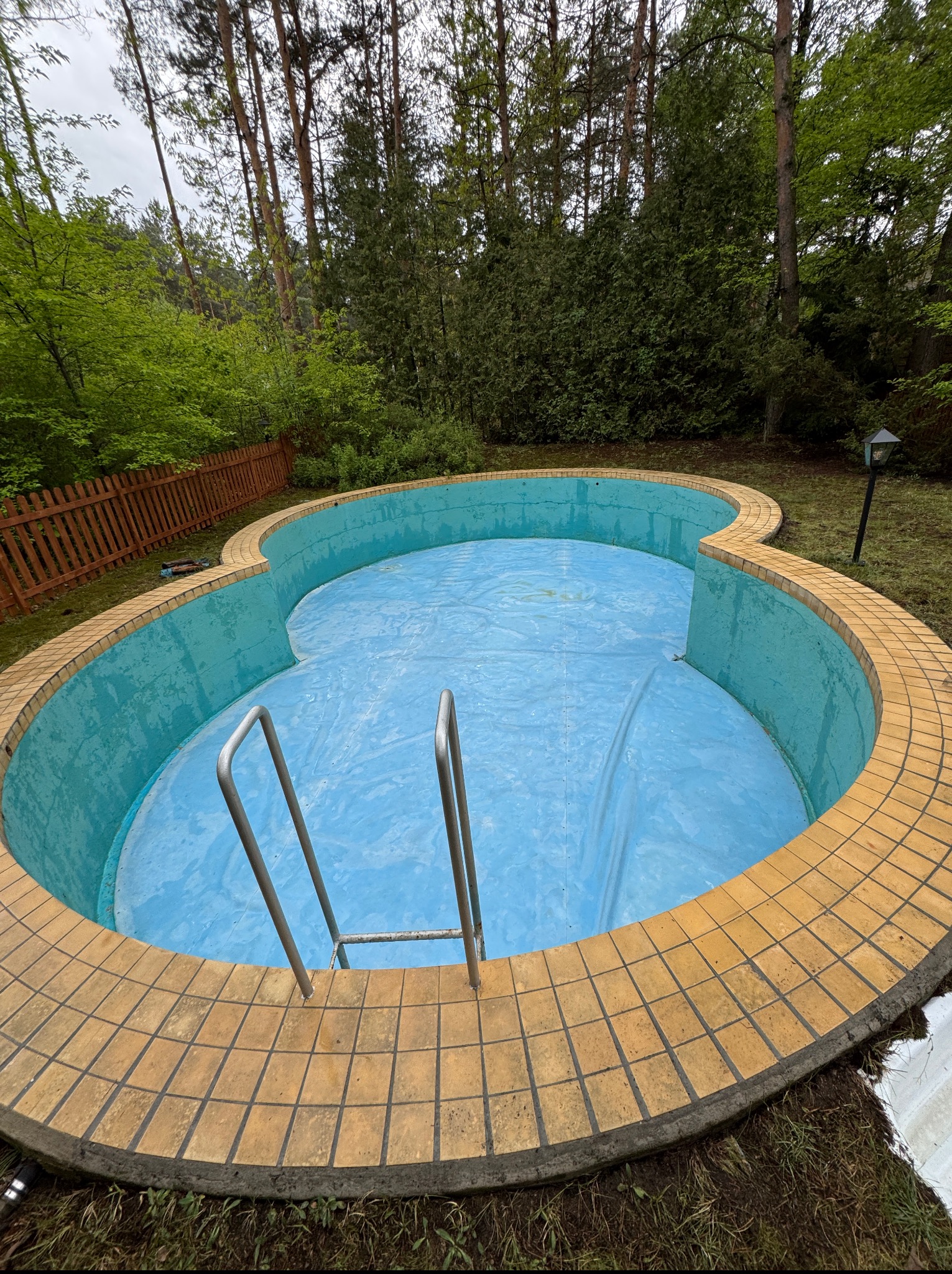 Nietypowy basen ogrodowy w kształcie koniczyny, zabezpieczony niebieską plandeką. Widoczne metalowe schodki, obrzeże z terakoty i otaczająca zieleń.