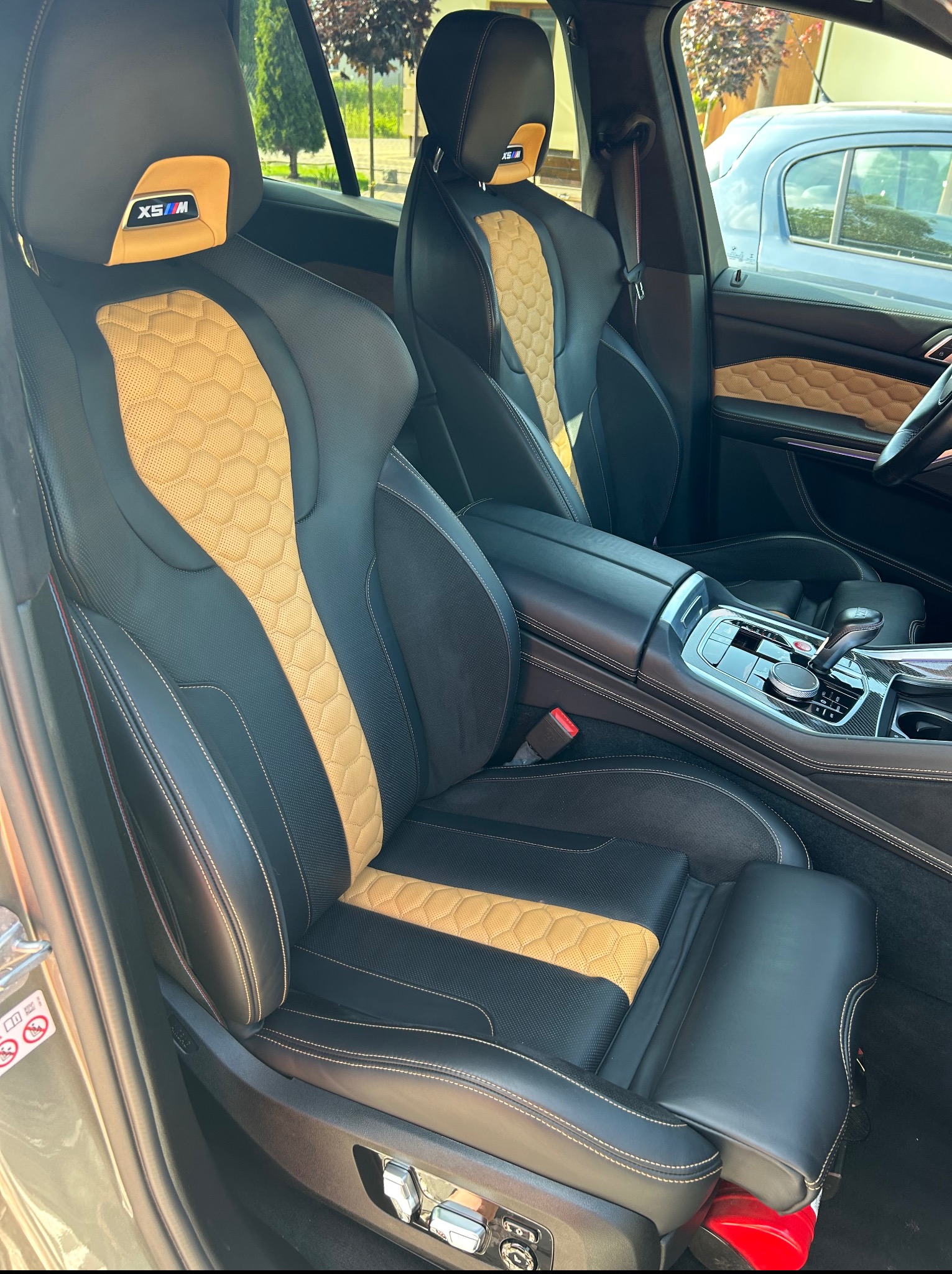 Wnętrze luksusowego samochodu z czarno-złotymi skórzanymi fotelami, widoczne detale przeszyć i konsola środkowa. Fotel kierowcy z logo X5M na zagłówku.