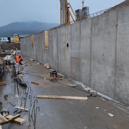 Szymon maciaszek - Wysoki żelbetowy mur oporowy w trakcie budowy, widoczny żuraw budowlany i elementy zbrojenia. Szary, pochmurny dzień. Widoczne otwory w murze.