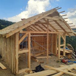 Szymon maciaszek - Drewniana konstrukcja altany w trakcie budowy w Rupniowie. Widoczne rusztowania, deski i narzędzia. W tle zalesione wzgórza i błękitne niebo z chmurami.