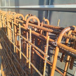 Szymon maciaszek - Zbrojenie fundamentów z bliska, z widocznymi detalami łączeń drutem i rdzawym nalotem, oświetlone bocznym światłem słonecznym na tle szarej ściany.