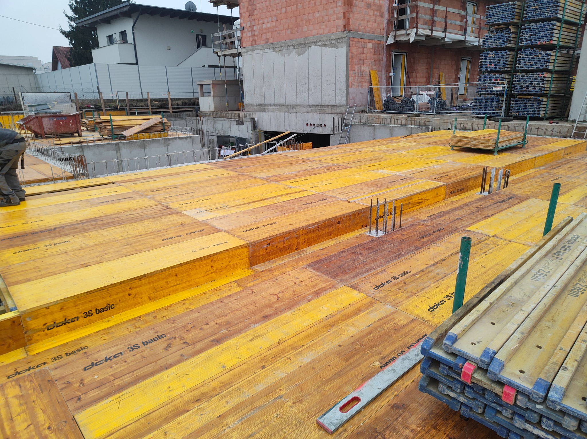 Żółte szalunki Doka 3S Basic tworzą platformę na budowie budynku żelbetowego. Widoczne zbrojenia, poziomica i stosy szalunków. W tle budynek w trakcie budowy.