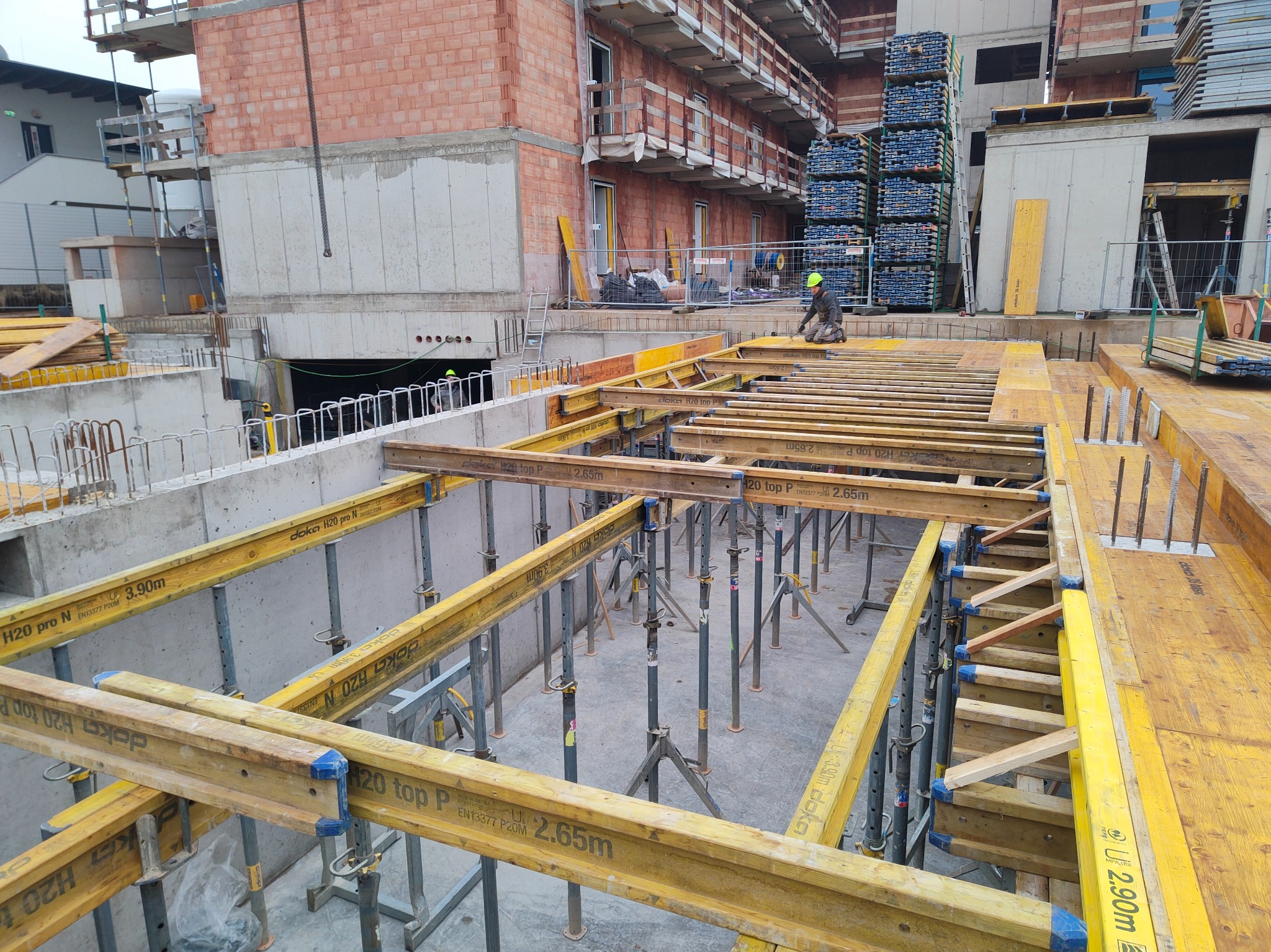 Szczegółowe ujęcie szalunku stropowego z żółtymi dźwigarami Doka H20 i stalowymi podporami na budowie budynku żelbetowego. Widoczny pracownik w kasku.