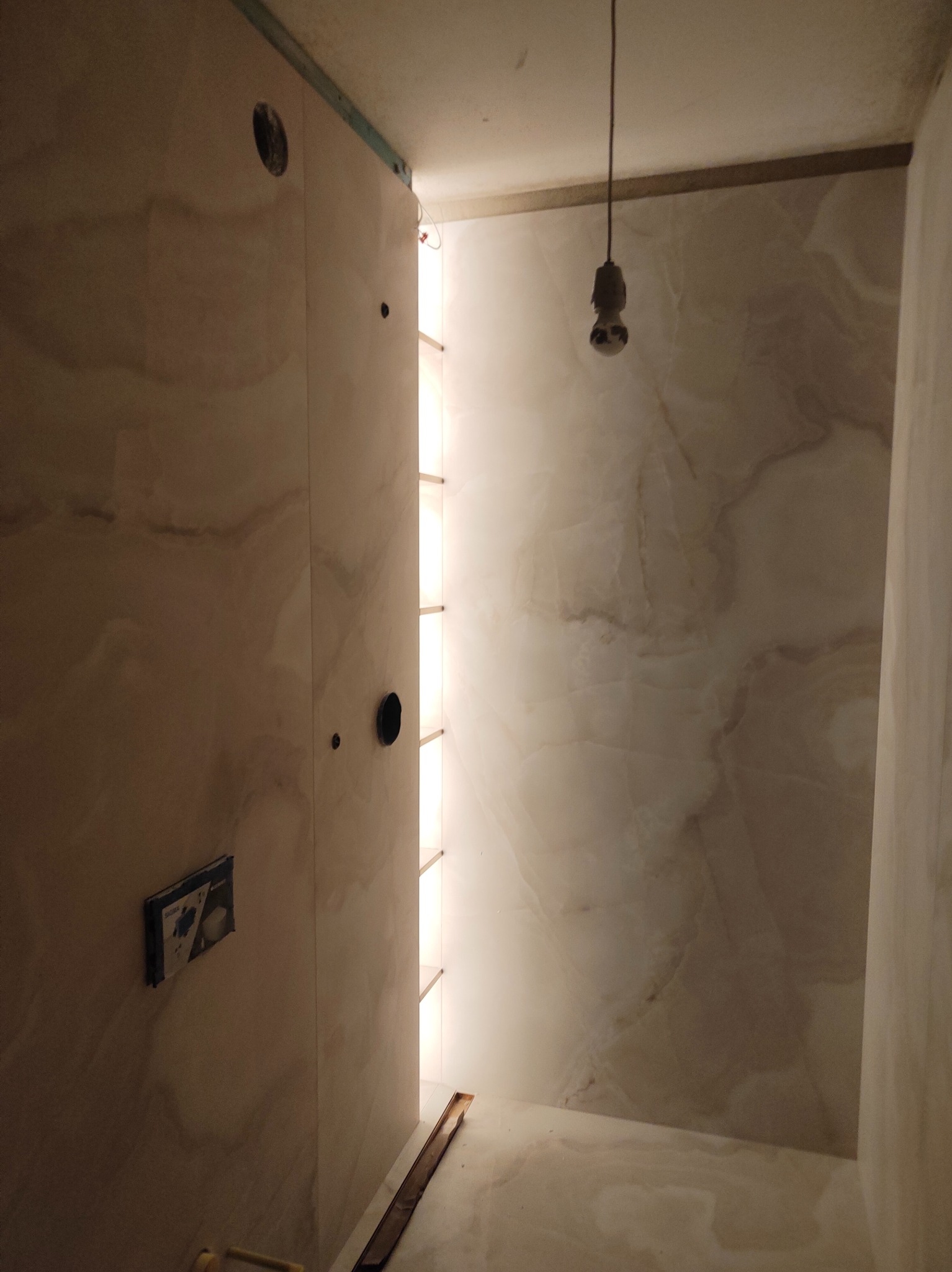 Wnętrze łazienki w trakcie wykańczania: marmurowe ściany, otwory na instalacje, podświetlane półki, surowa żarówka. Widok z dołu, podkreślający przestrzeń.