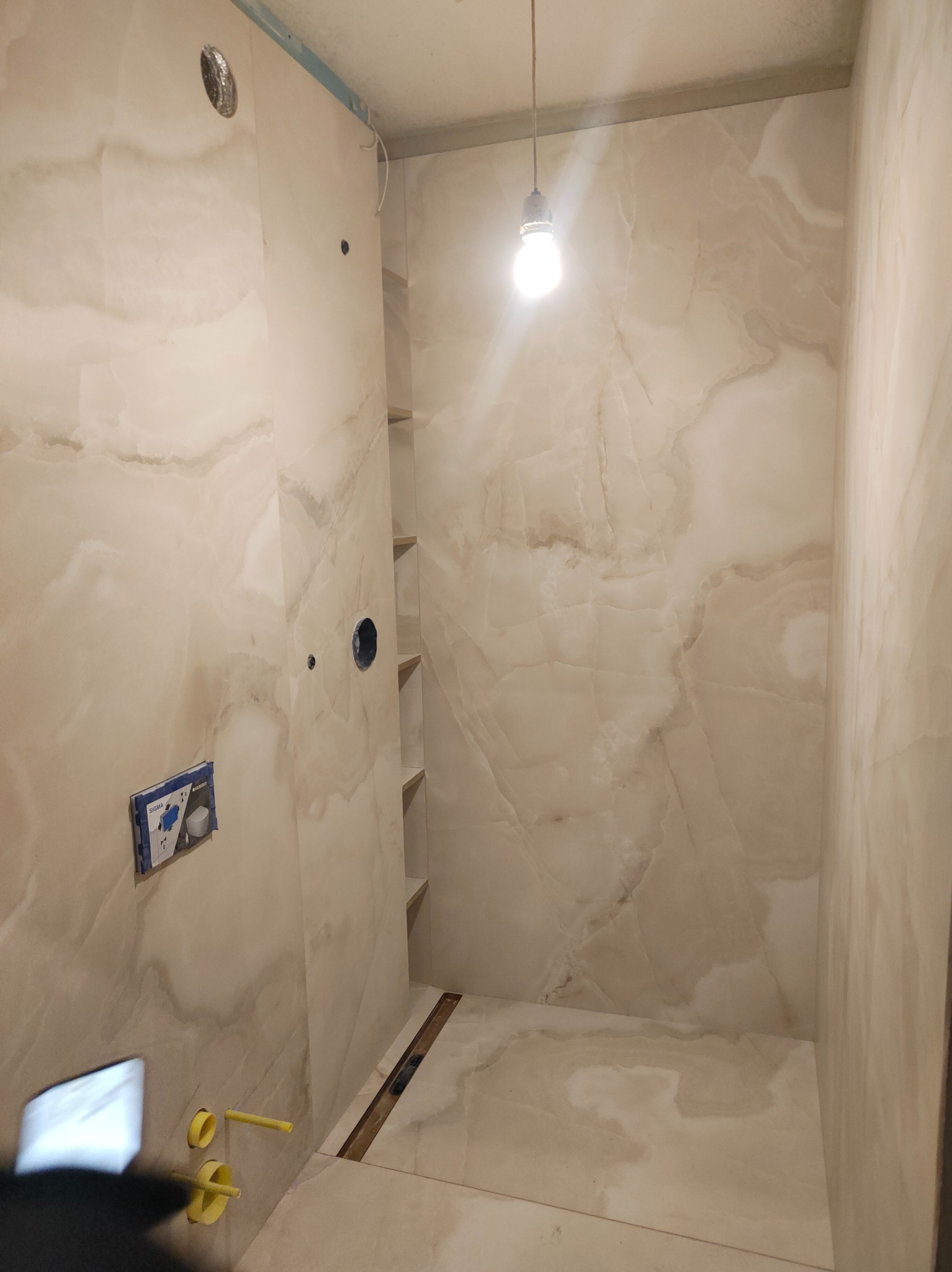 Wykończona łazienka z beżowymi płytkami imitującymi marmur, widoczne instalacje i wnęki półkowe. Oświetlenie żarówką. Ujęcie z dołu, podkreślające przestrzeń.