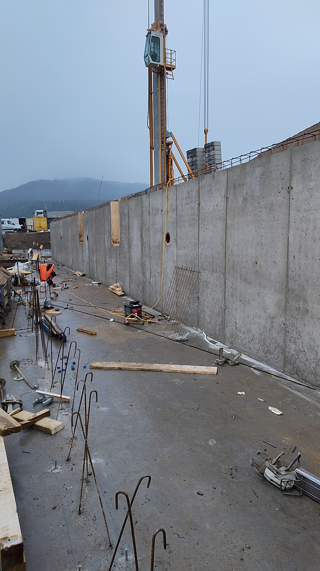 Wysoki żelbetowy mur oporowy w trakcie budowy, widoczny żuraw budowlany i elementy zbrojenia. Szary, pochmurny dzień. Widoczne otwory w murze.