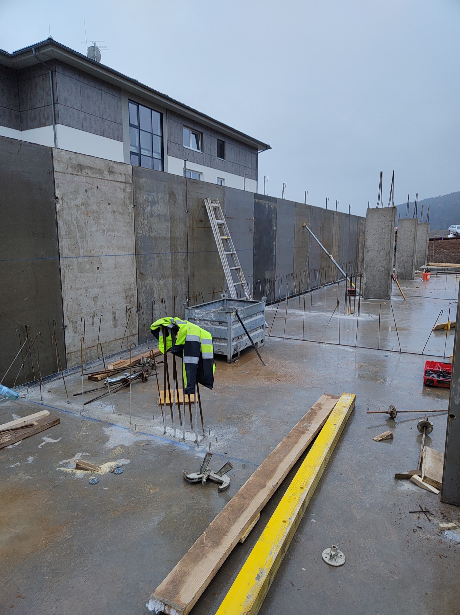 Szare szalunki pionowe zbrojone stalą, przygotowane do zalania betonem, na tle dwukondygnacyjnego budynku w pochmurny dzień. Widoczne narzędzia i odzież robocza.