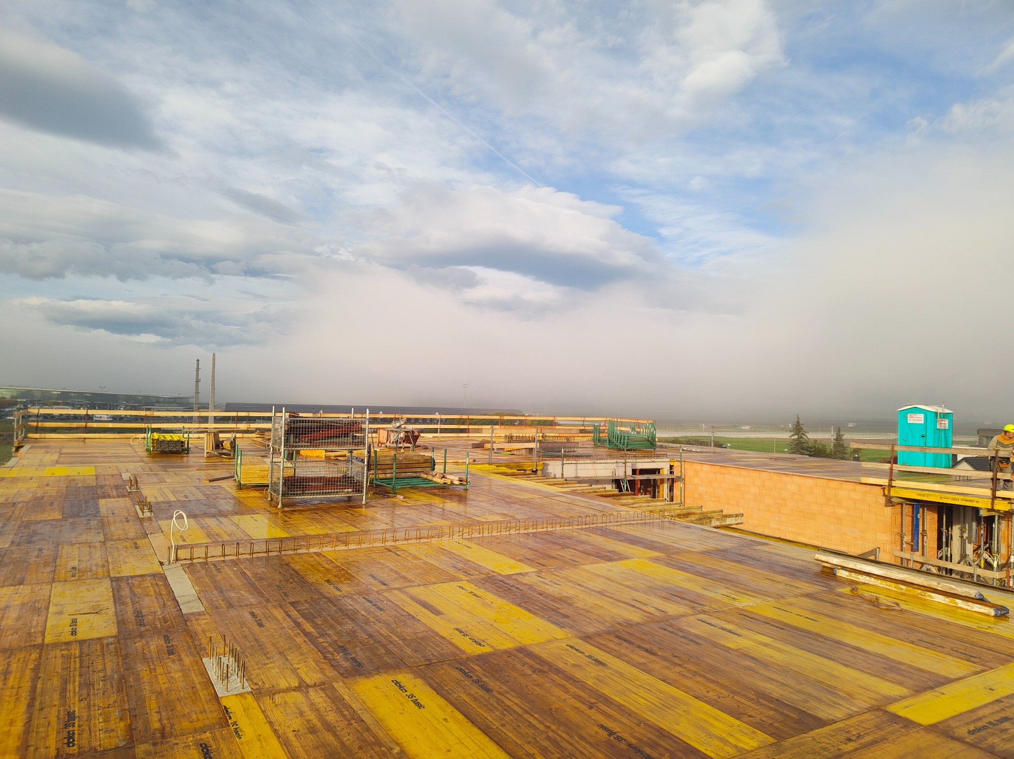 Żółte szalunki na budowie budynku żelbetowego, widok z góry na konstrukcję w trakcie realizacji, z boku widoczna toaleta przenośna i pracownik w kasku.