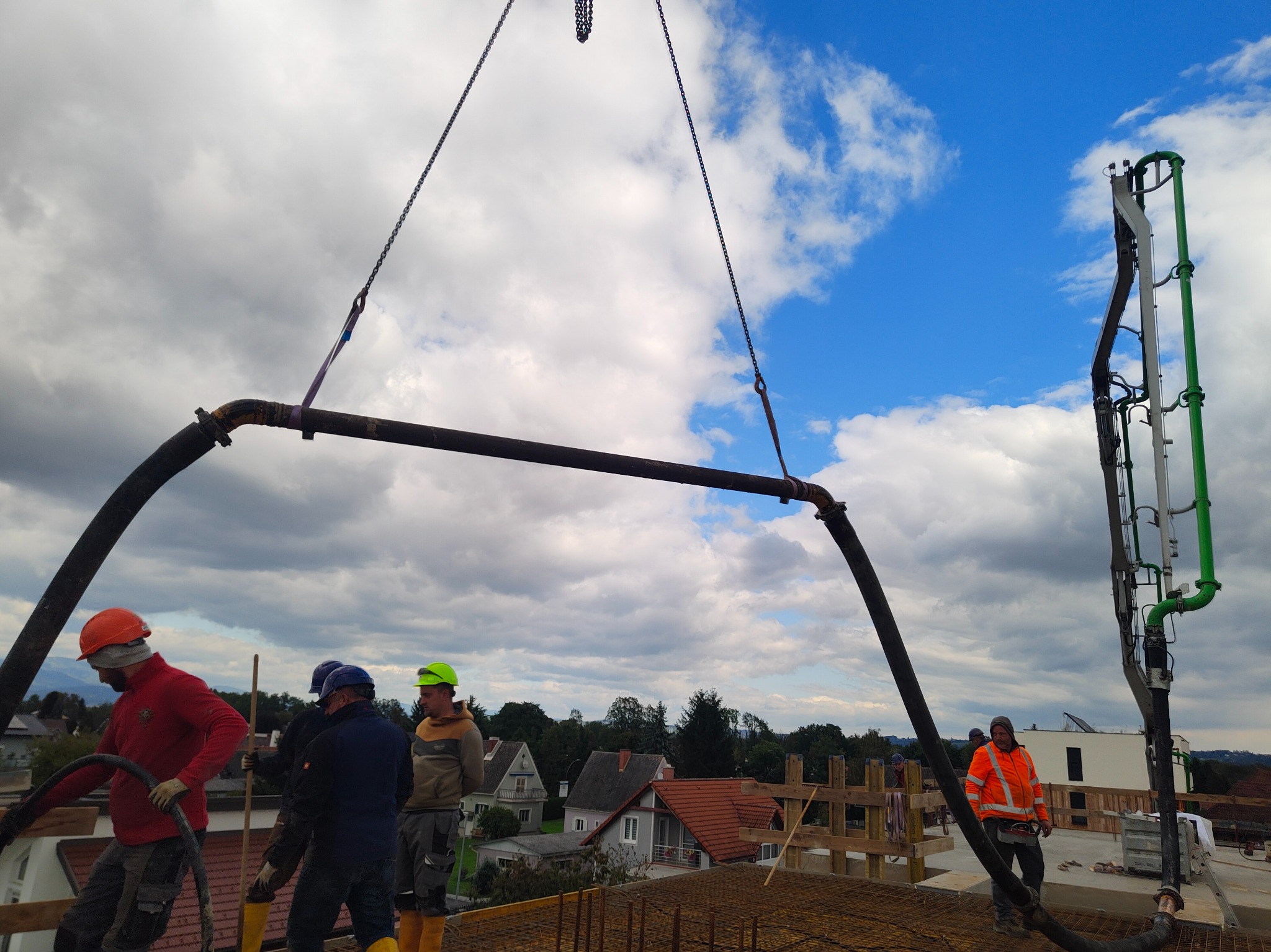 Betonowanie stropu budynku z użyciem pompy do betonu. Widoczni pracownicy w kaskach i roboczych ubraniach. W tle zabudowa jednorodzinna i pochmurne niebo.