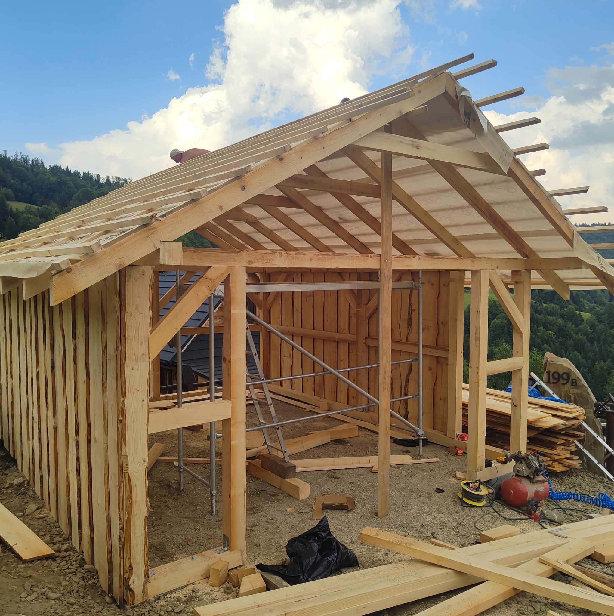 Drewniana konstrukcja altany w trakcie budowy w Rupniowie. Widoczne rusztowania, deski i narzędzia. W tle zalesione wzgórza i błękitne niebo z chmurami.