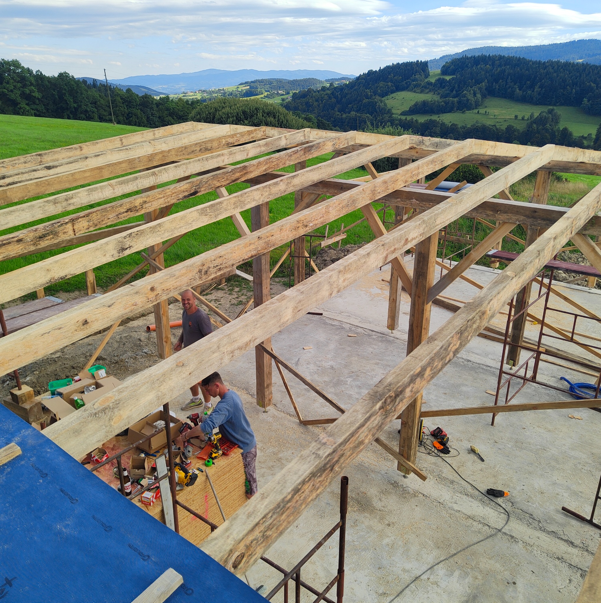 Drewniana konstrukcja dachu w trakcie budowy, widok z góry na robotników i narzędzia, w tle zielone wzgórza i błękitne niebo. Elementy rusztowania.