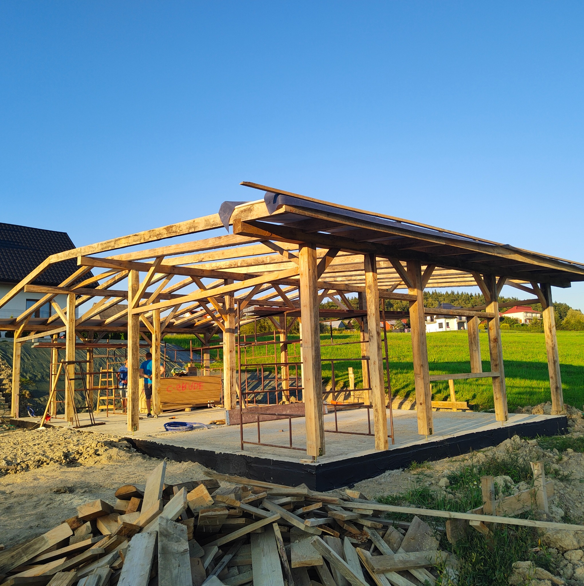 Drewniana konstrukcja altany w trakcie budowy, widoczne elementy szkieletu i fundamenty. W tle zielona trawa i błękitne niebo.