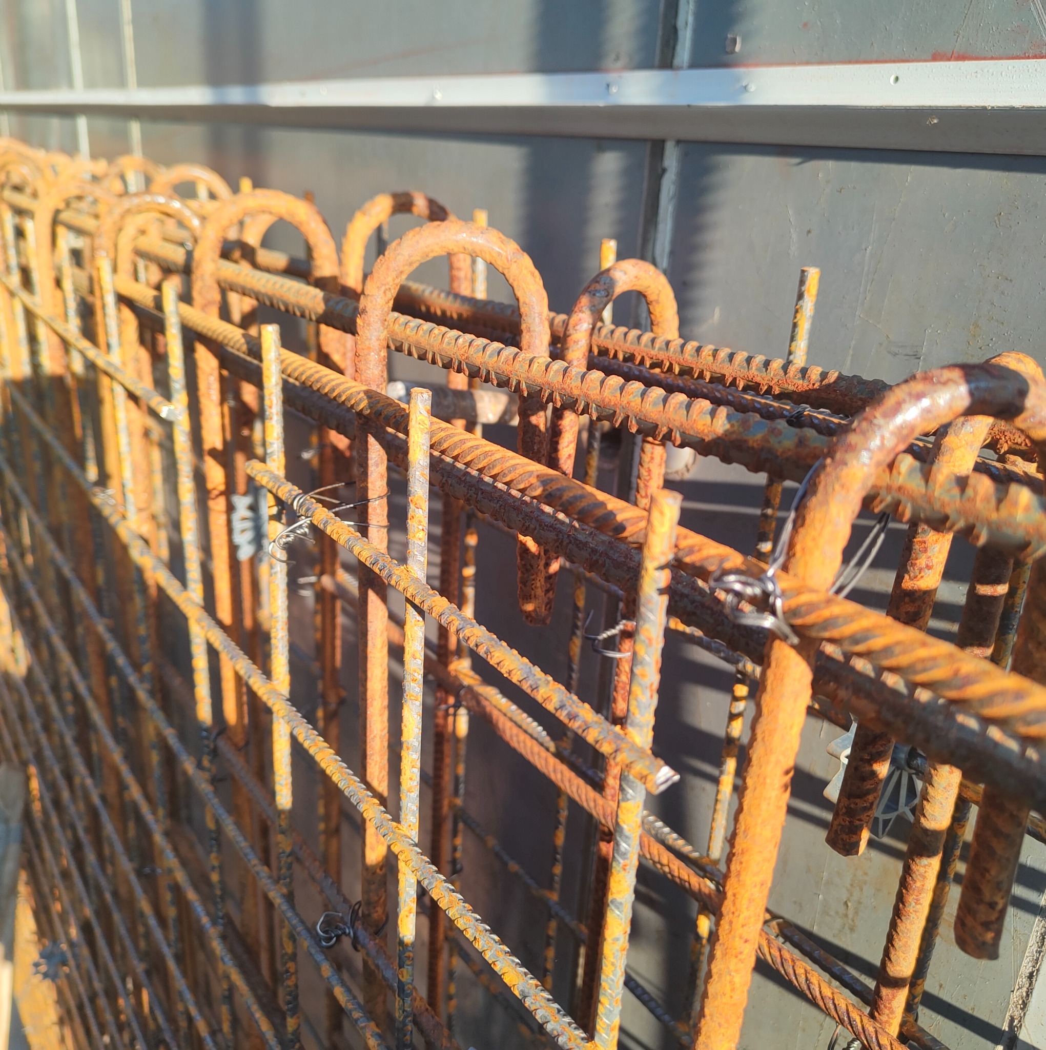 Zbrojenie fundamentów z bliska, z widocznymi detalami łączeń drutem i rdzawym nalotem, oświetlone bocznym światłem słonecznym na tle szarej ściany.