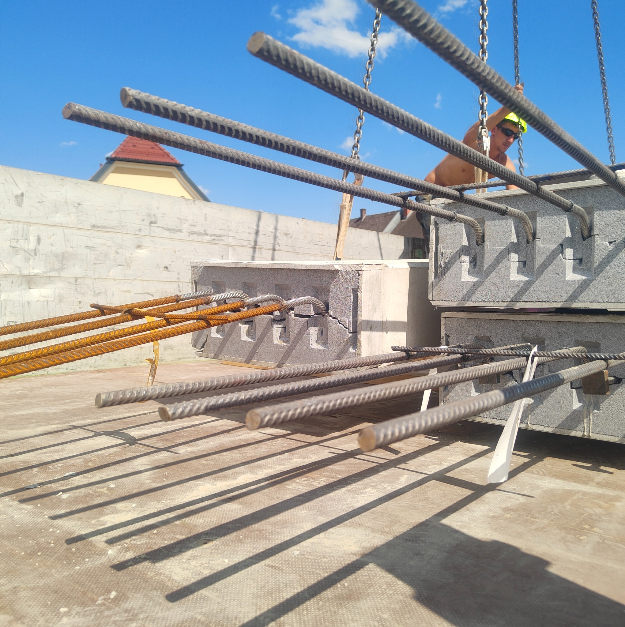 Zbrojenie stropu Teriva z żelbetowych belek i pustaków szalunkowych, widoczne wystające pręty i pracownik w kasku. Słoneczny dzień, budowa w toku.