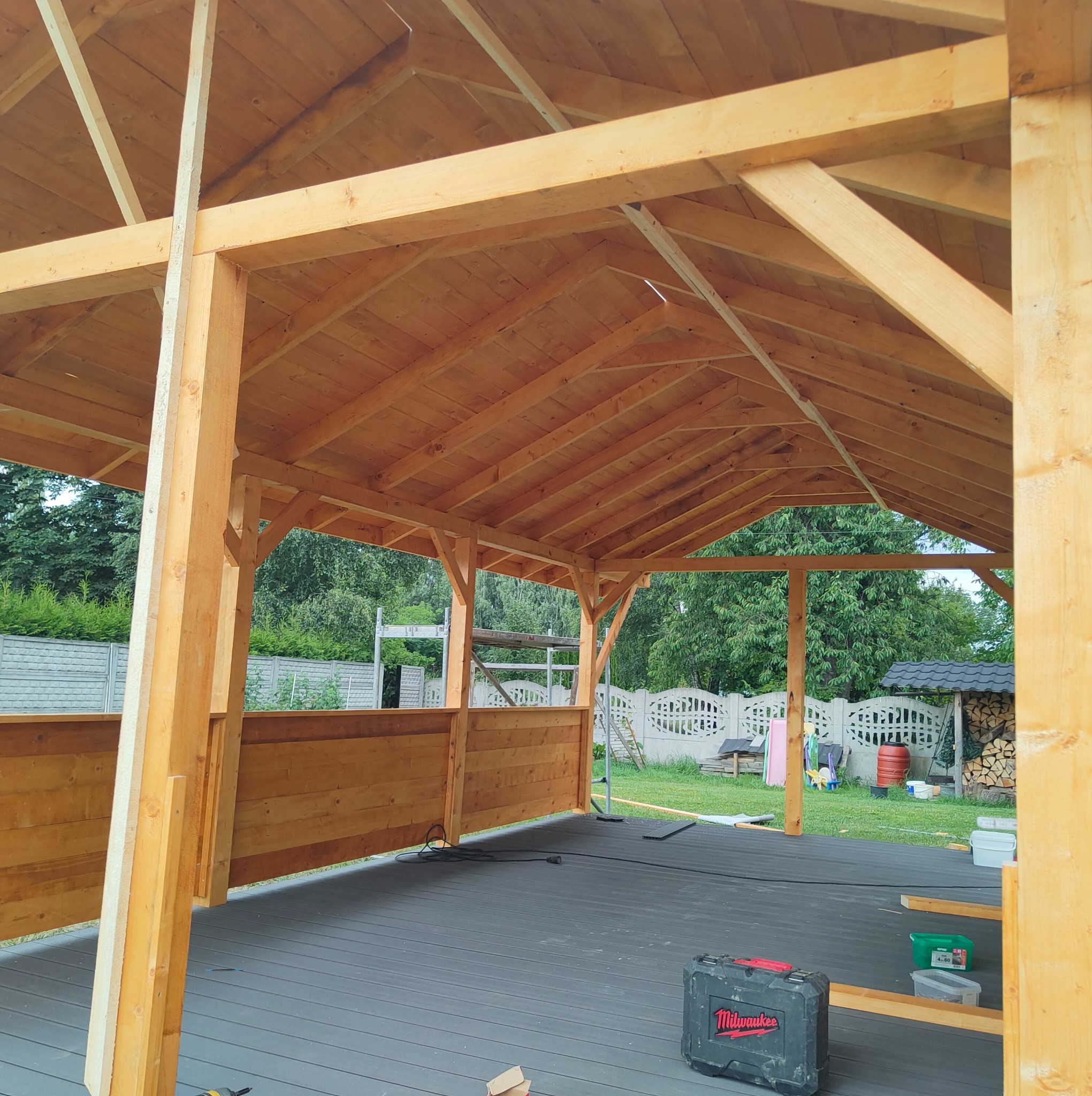 Drewniana konstrukcja altany w trakcie budowy, widoczne elementy szkieletu i podłogi z desek kompozytowych. W tle zieleń ogrodu i ogrodzenie.