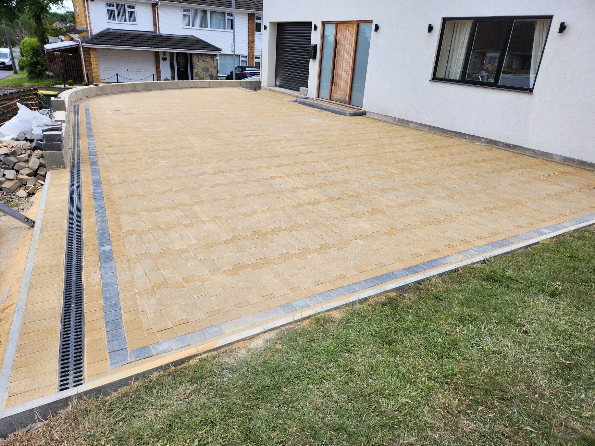 Nowoczesny podjazd z kostki brukowej w odcieniach beżu i szarości, z eleganckim systemem odprowadzania wody. Dom z białym tynkiem i czarnymi ramami okiennymi w tle.