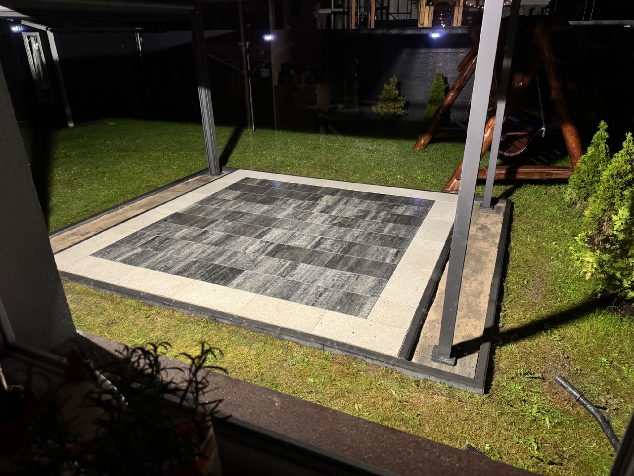 Nowoczesny taras z kostki brukowej w odcieniach szarości i beżu, otoczony zielonym trawnikiem i elementami architektury ogrodowej. Całość oświetlona w nocy.