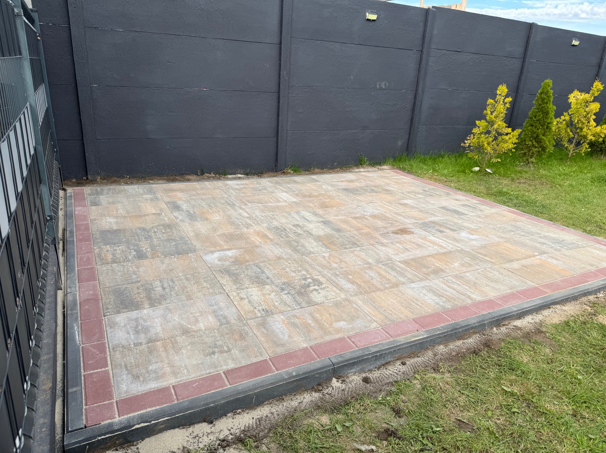 Ułożona kostka brukowa w odcieniach szarości i beżu, obramowana czerwoną kostką. W tle ciemnoszary, betonowy płot i zieleń trawy. Widok na gotową nawierzchnię.