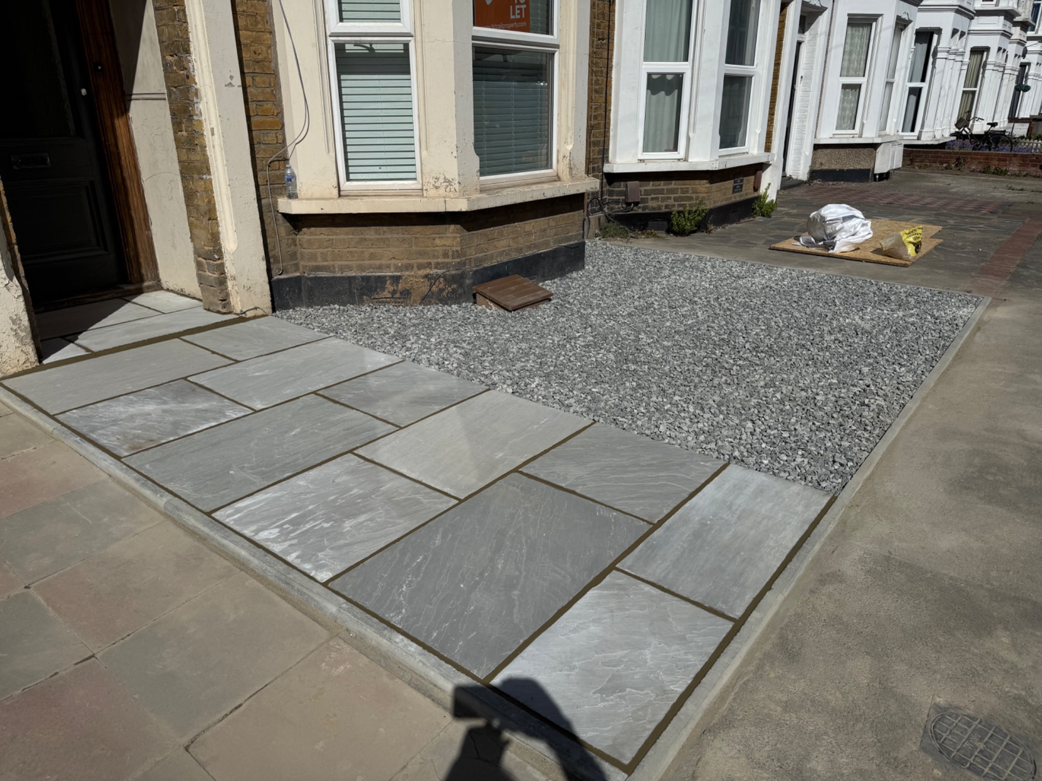 Nowa nawierzchnia z szarych płyt i kamieni przed domem. Widoczne okna i drzwi wejściowe. Estetyczne wykończenie, równo ułożone elementy, schludny wygląd.