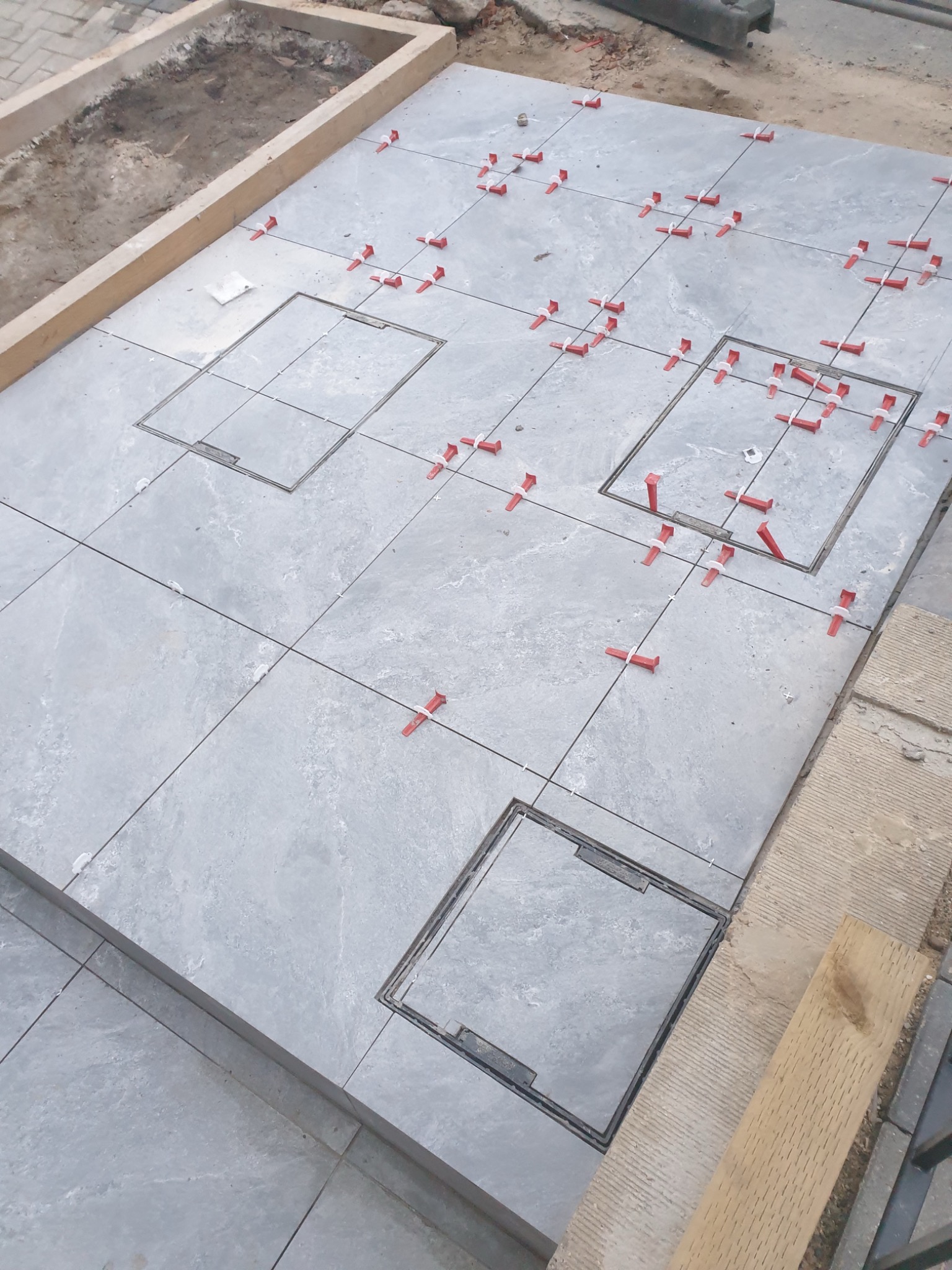 Szare płytki z widocznymi włazami i czerwonymi krzyżykami dystansowymi, ułożone na zewnątrz, otoczone drewnianą ramą, w trakcie prac wykończeniowych.