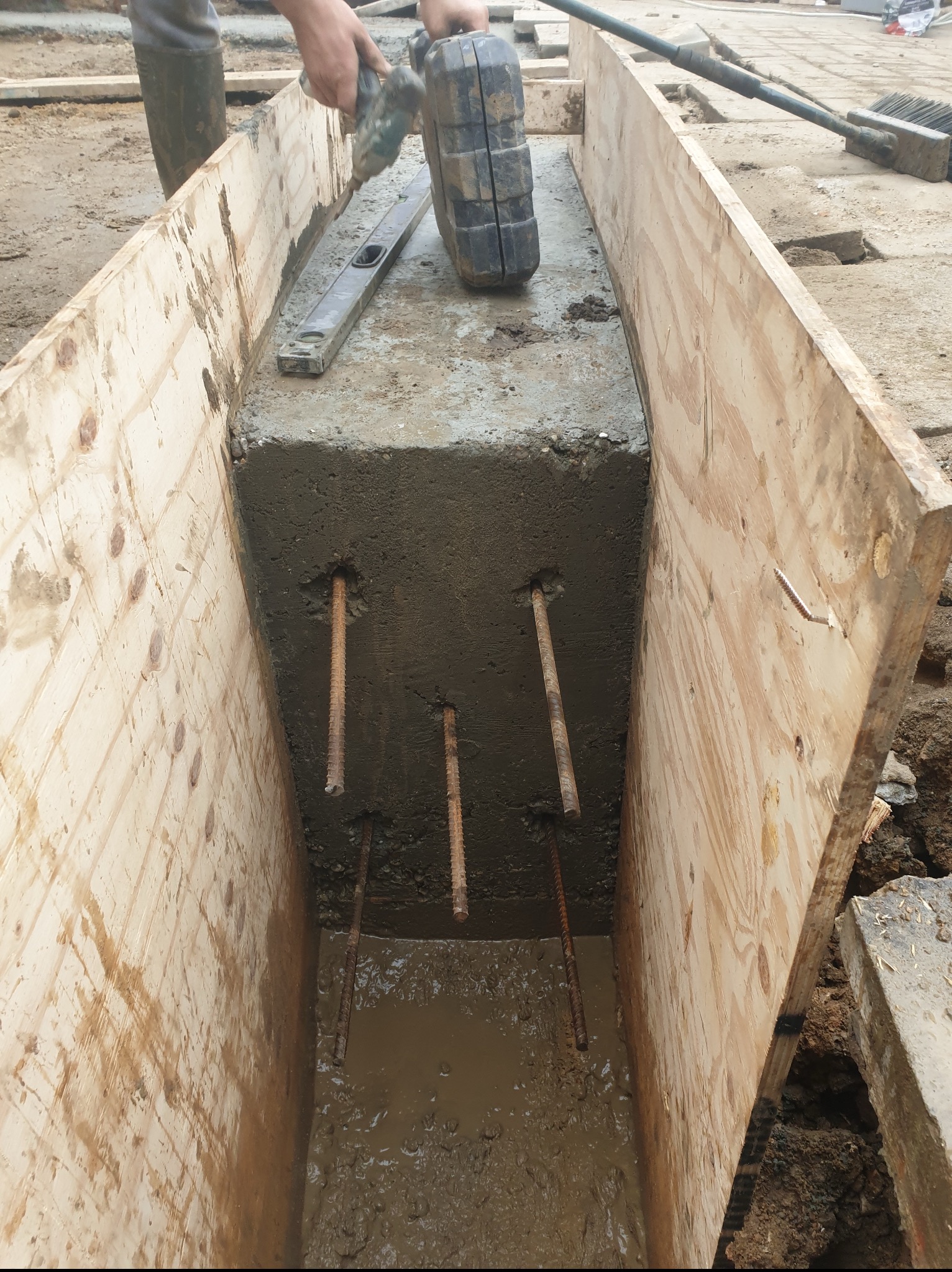 Zalewanie betonem fundamentu pod budynek, widoczne zbrojenie i drewniane szalunki. Prace ziemne w toku, poziomica i narzędzia obok.