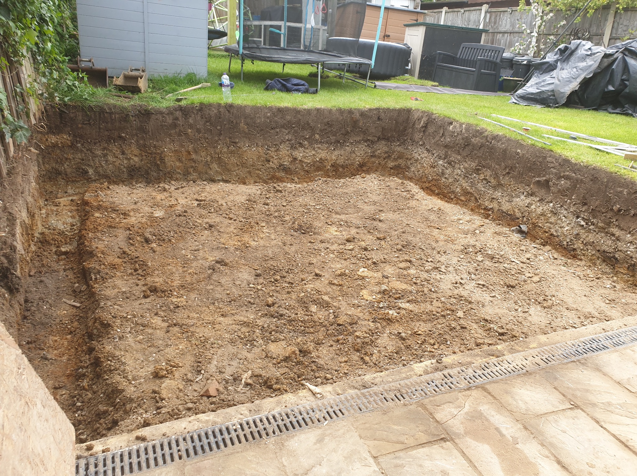 Wykopany dół w ziemi, prawdopodobnie pod basen lub oczko wodne, z widocznymi warstwami gleby i elementami krajobrazu w tle.