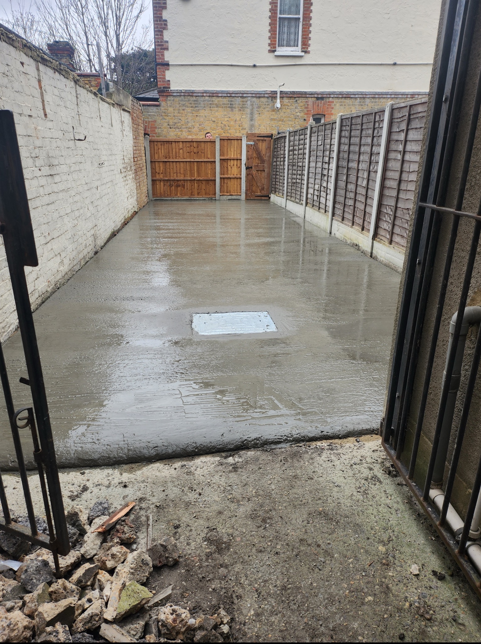 Wylany beton na podwórku z kratką ściekową, otoczony murem z cegły i drewnianym płotem. Fragment otwartej, metalowej bramy w kadrze.