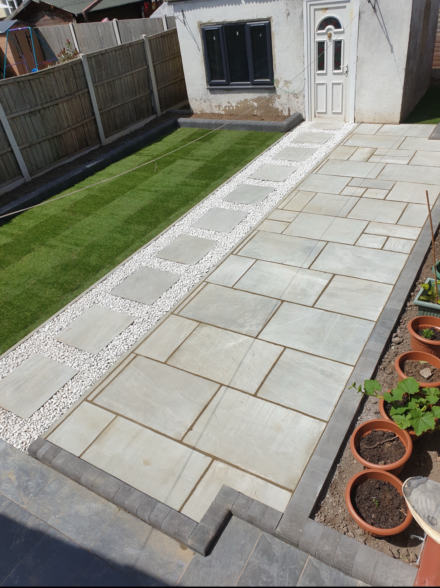 Ułożona kostka brukowa w ogrodzie z białym żwirem i trawą, prowadząca do budynku z białymi drzwiami i oknem. Widok z góry na nową nawierzchnię.