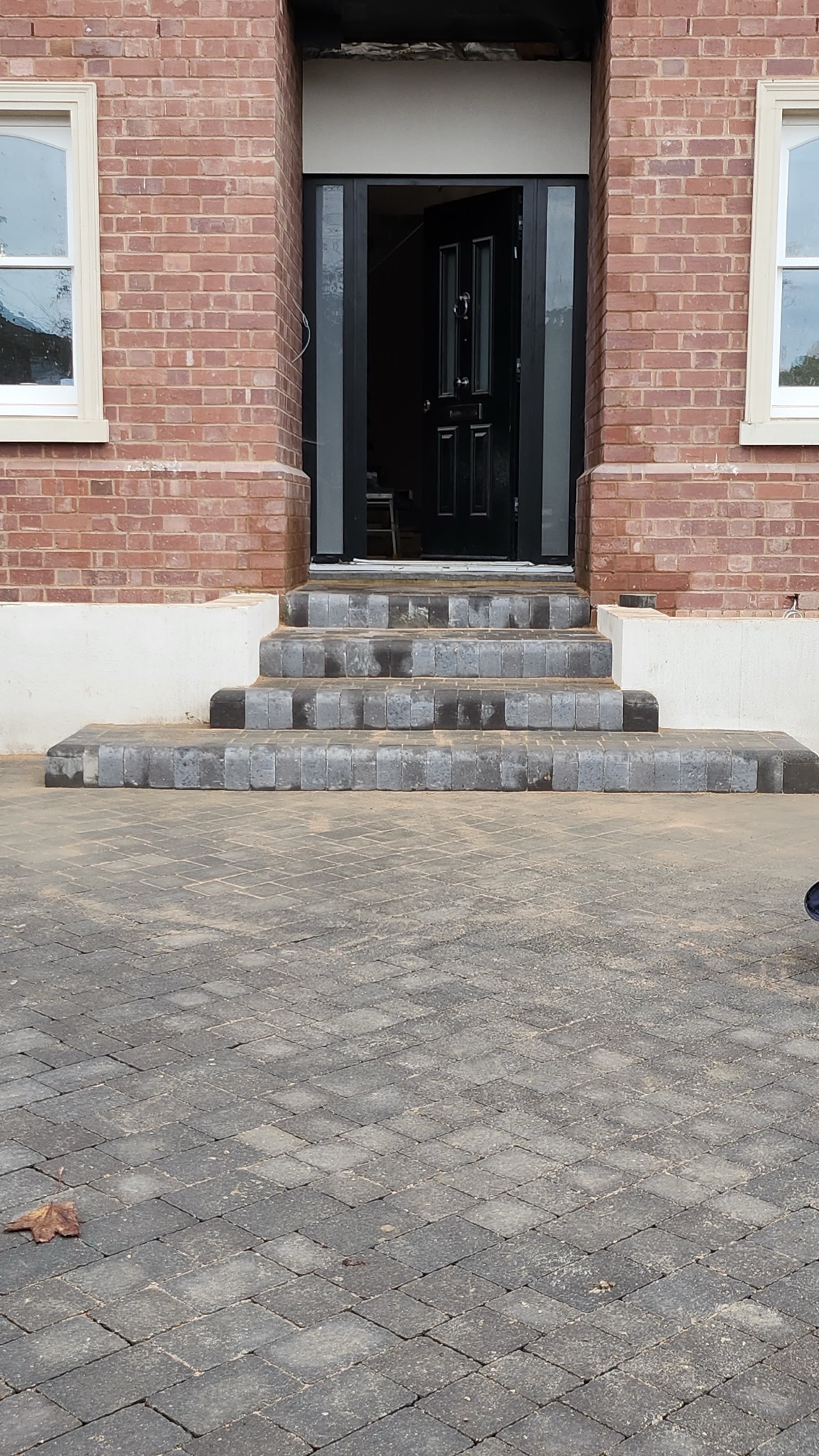 Ułożona kostka brukowa prowadzi do wejścia domu z cegły. Czterostopniowe schody z kostki brukowej. Widoczny fragment liścia na kostce brukowej.