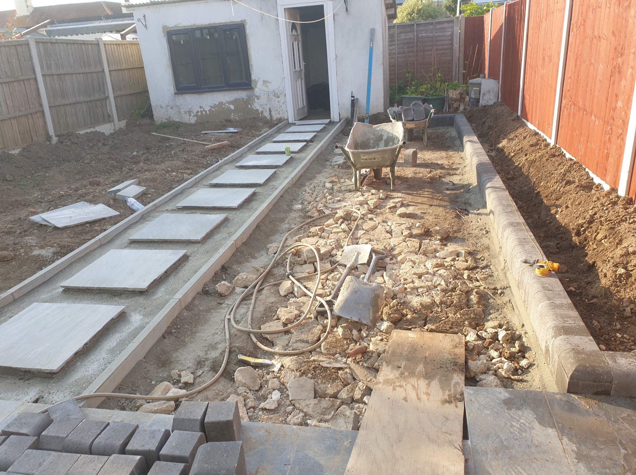 Kształtowanie ścieżki z płyt i obrzeży z kostki brukowej w ogrodzie, widoczne narzędzia i materiały budowlane. Etap prac ziemnych i brukowania w toku.