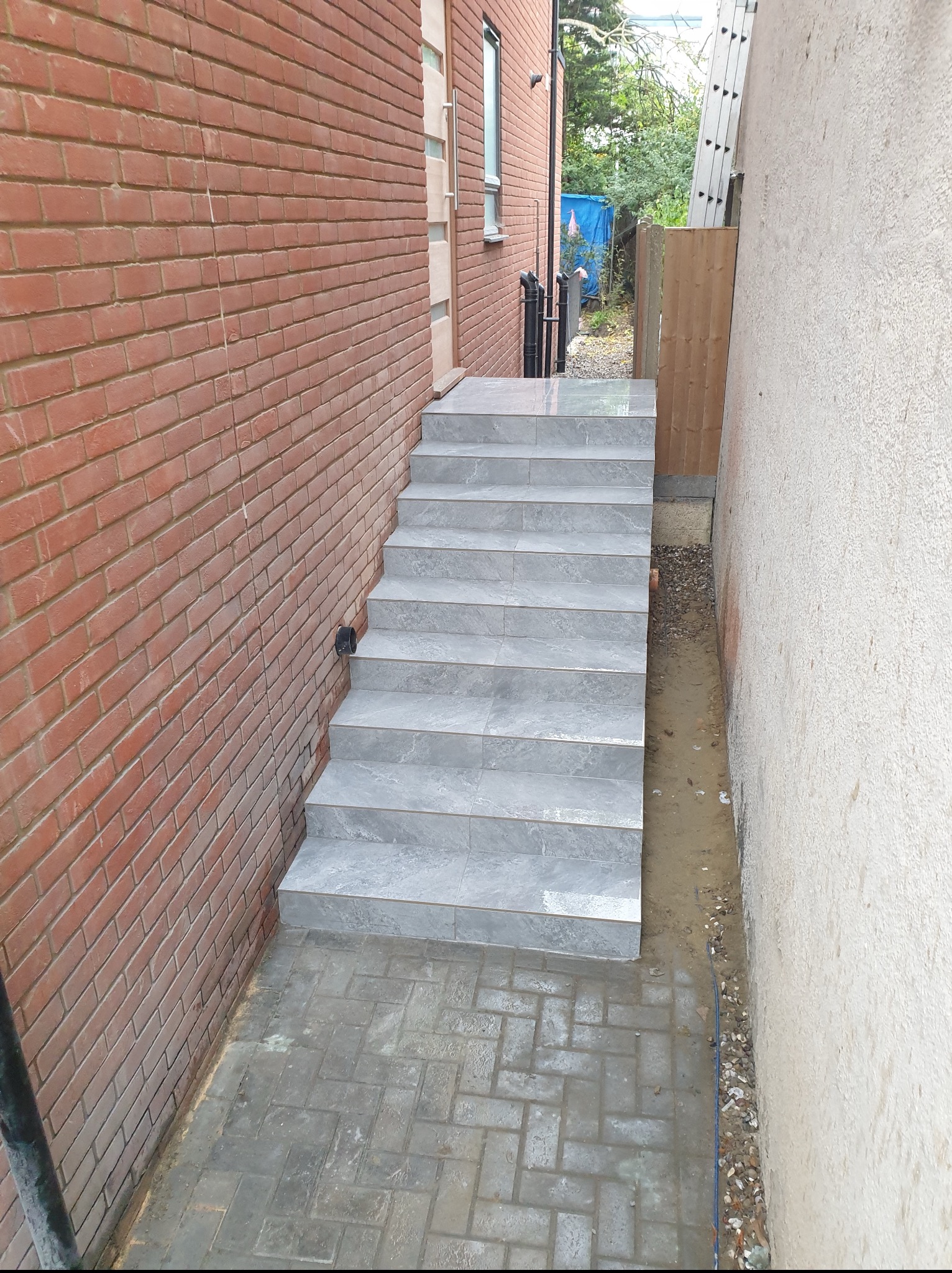Zewnętrzne schody z płytek w szarej tonacji, prowadzące z chodnika z kostki brukowej. Ciasna przestrzeń między budynkami z cegły i otynkowaną ścianą.