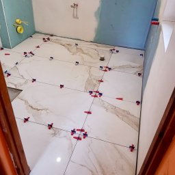 DABSTER DARIUSZ BACHULA - Łazienka w trakcie prac: białe płytki imitujące marmur, system poziomowania płytek, częściowo wykończone ściany z instalacją hydrauliczną. Widok z progu.