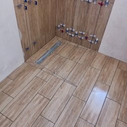 DABSTER DARIUSZ BACHULA - Kafelkowanie łazienki w trakcie: brązowe płytki imitujące drewno na ścianach i podłodze, system poziomowania płytek. Widoczny odpływ liniowy.