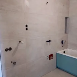 Łazienka w trakcie wykańczania: beżowe płytki na ścianach, przygotowane punkty instalacyjne, zabudowa wanny z płyty GK. Widoczny etap prac glazurniczych i instalacyjnych.