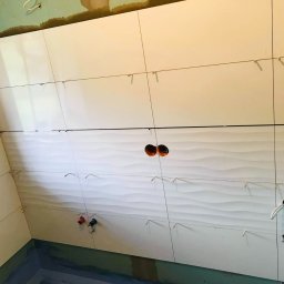 DABSTER DARIUSZ BACHULA - Ściana w trakcie układania białych płytek z falistym wzorem, widoczne otwory na instalacje i krzyżyki dystansowe. Etap prac w łazience.