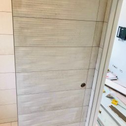 DABSTER DARIUSZ BACHULA - Ściana wyłożona beżowymi, poziomymi płytkami imitującymi drewno, z widocznymi otworami i fragmentem łazienki w tle, ukazana z perspektywy bliskiej.