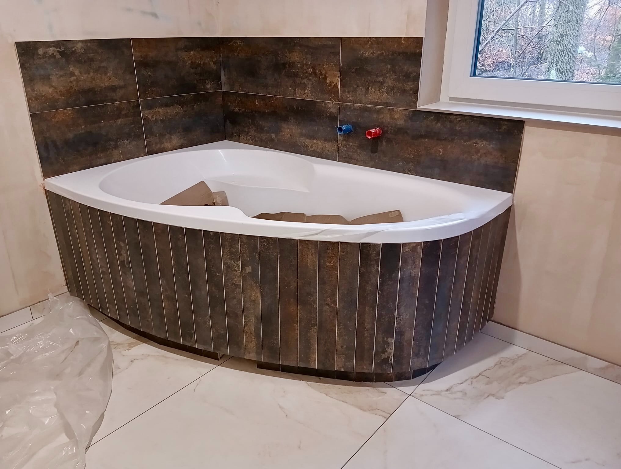 Narożna wanna obudowana ciemnymi, pionowo ułożonymi płytkami. Na ścianie brązowe płytki imitujące rdzę, biała marmurowa podłoga. Widoczne przyłącza wodne.