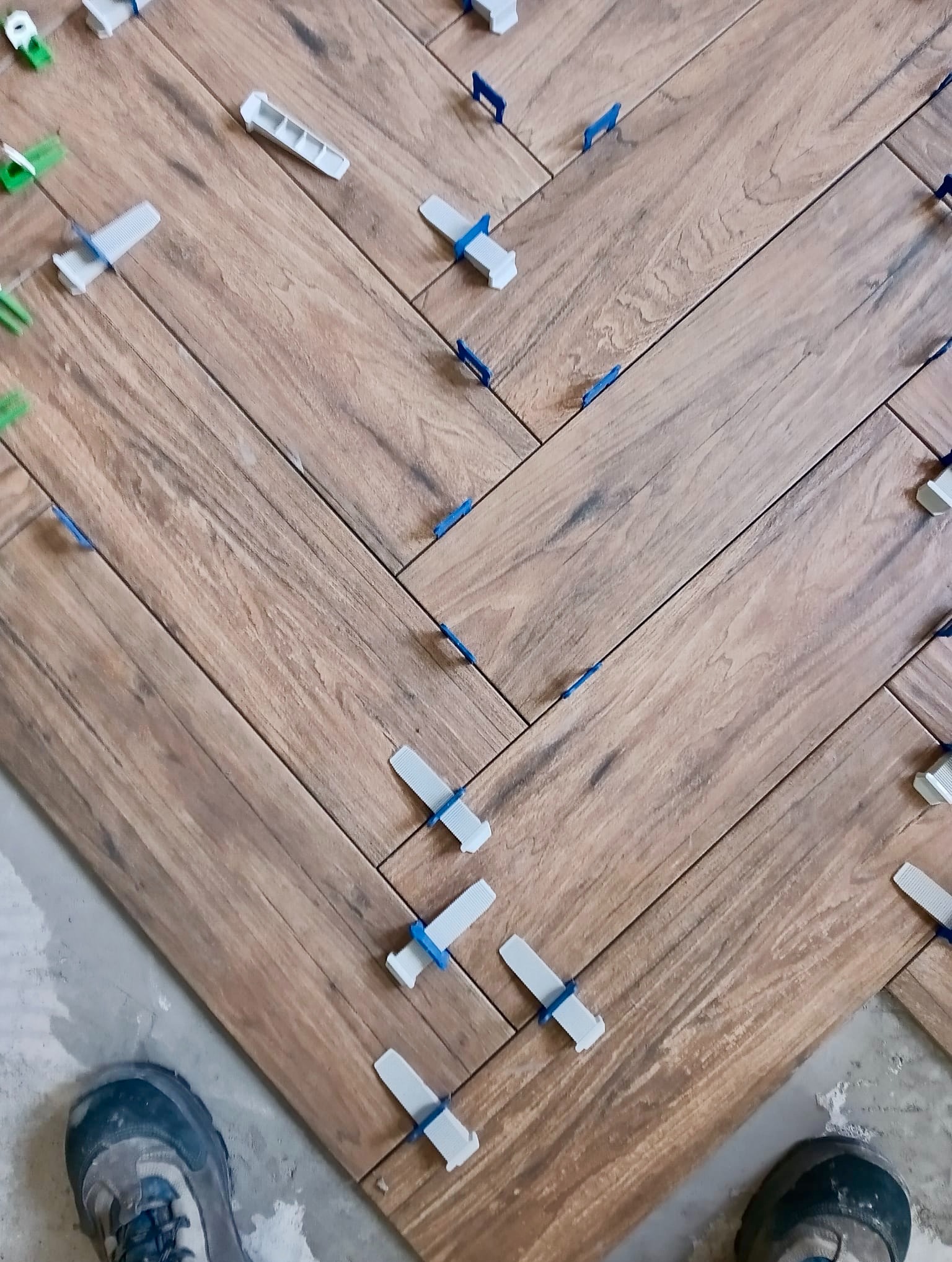 Układanie drewnopodobnych płytek w jodełkę z użyciem klipsów poziomujących. Widoczne buty robocze w dolnej części kadru.