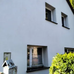 Biała elewacja domu z trzema oknami, widok z dołu. Z prawej strony krzew, z lewej skrzynka elektryczna. Świeżo pomalowana fasada z widoczną strukturą.