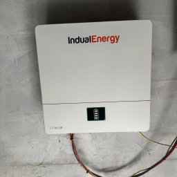 Entermic sp. z o. o. - Biały magazyn energii IndualEnergy zamontowany na szarej, surowej ścianie. Widoczne kable i wskaźnik naładowania baterii.