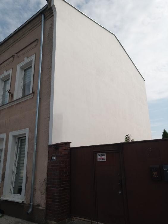 Ocieplona, biała ściana budynku z widocznym fragmentem elewacji przed ociepleniem. Rynna pionowa z boku. Brama wjazdowa z tabliczką informacyjną.