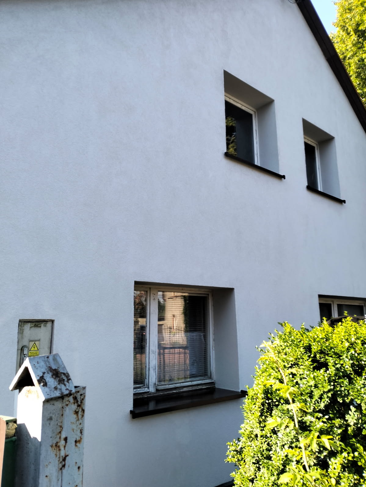 Biała elewacja domu z trzema oknami, widok z dołu. Z prawej strony krzew, z lewej skrzynka elektryczna. Świeżo pomalowana fasada z widoczną strukturą.