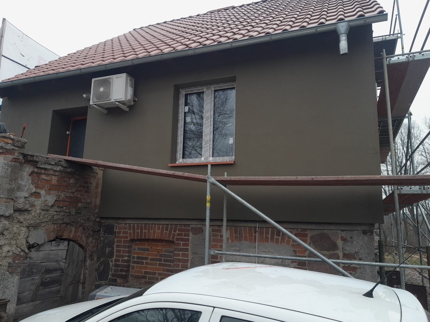 Ocieplony budynek z nową elewacją w odcieniach szarości, z zamontowanym klimatyzatorem i oknem. Widoczne rusztowanie i fragment ceglanego muru. Częściowo widoczny samochód.