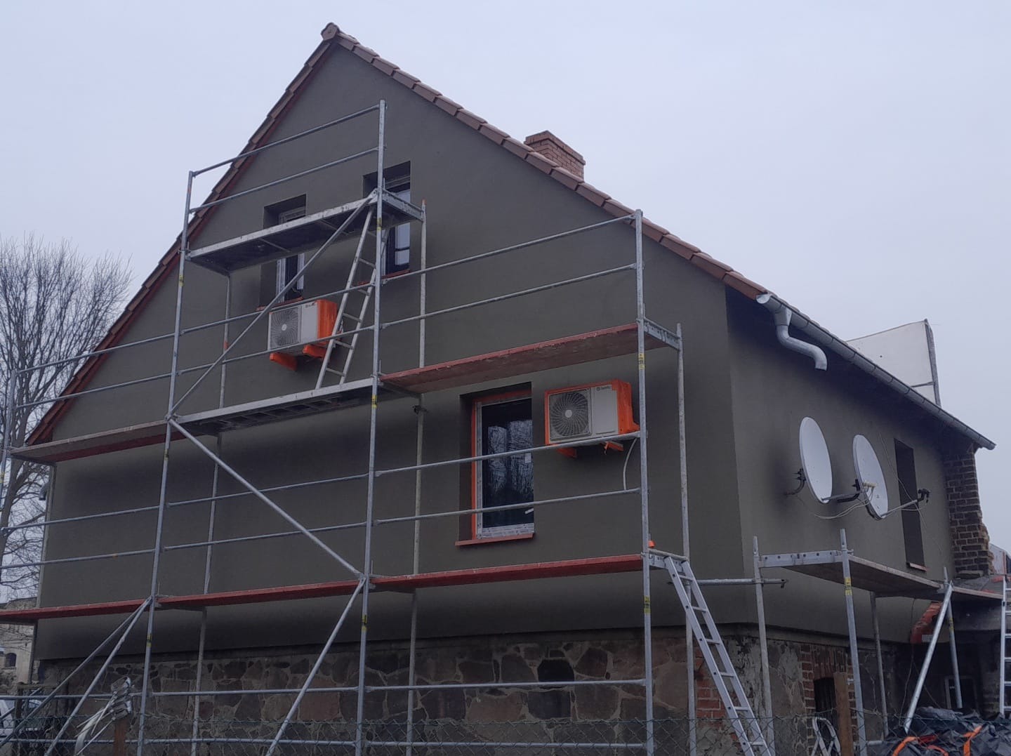 Dom w trakcie prac elewacyjnych, z widocznymi rusztowaniami, oknami z pomarańczowymi ramami i klimatyzatorami na ścianie. Ściany w kolorze szaro-zielonym.