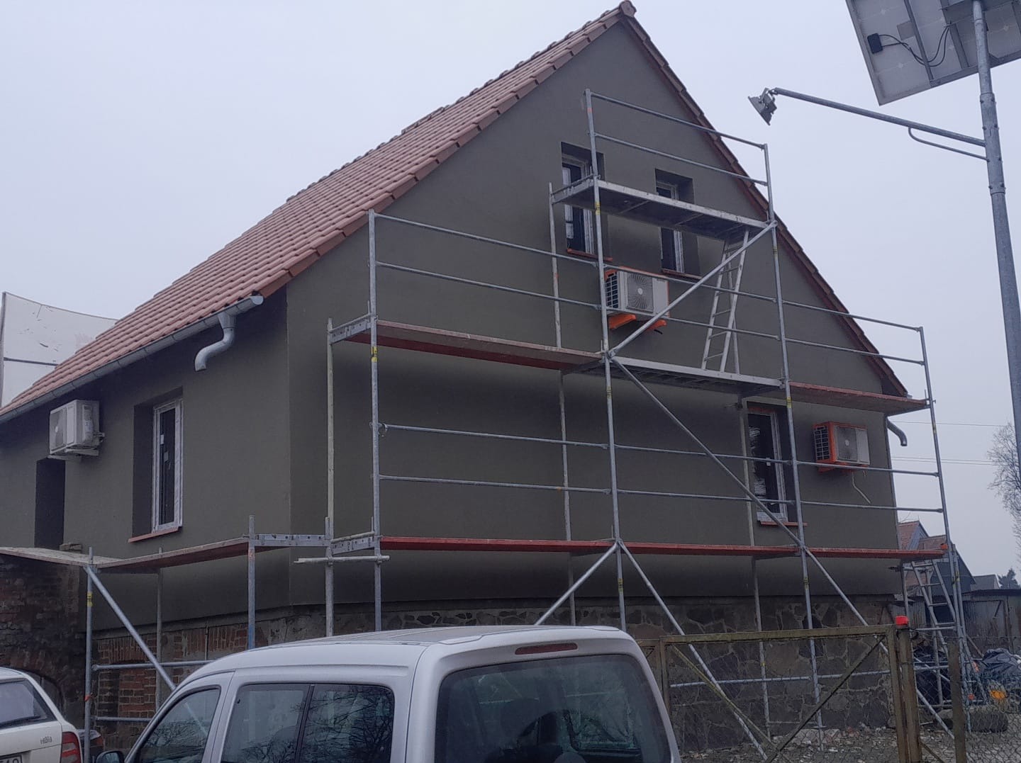 Dom po ociepleniu, z szarym tynkiem i nową stolarką okienną, otoczony rusztowaniem. Widoczne jednostki klimatyzacji na elewacji i fragment samochodu w tle.
